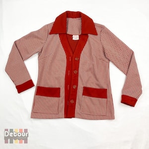 May include: A red and white patterned cardigan with a V-neck, long sleeves, and a button-up front. The cardigan has red trim around the collar, cuffs, and pockets.
