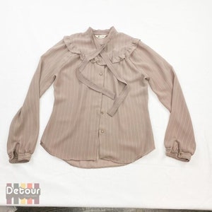 May include: A pale brown long-sleeved blouse with vertical stripes. The blouse has a ruffled collar, a tie at the neckline, and button closures down the front. The sleeves are slightly puffed at the shoulders and have button cuffs. The blouse is laid flat on a white surface.