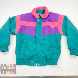 May include: A vintage colour-blocked jacket in teal, purple, and pink. The jacket features a high collar, a button-up front, and elastic cuffs and waist. The jacket has a small pocket on the chest and a small logo on the bottom. The jacket is from the brand Detour.