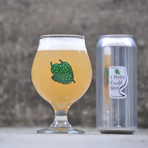May include: A glass of light-colored beer in a stemmed glass with a green hop sticker. A silver can of beer with a sticker that says "I Prefer Craft Beer" is next to the glass.