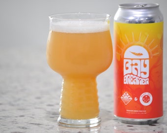 Könnte beinhalten: Eine Dose Bay Dreamer Session India Pale Ale mit einem Sonnenuntergangs-Motiv und dem Text "Bay Dreamer" in Weiß. Die Dose steht neben einem Glas des Bieres, das eine trübe orange Farbe hat.