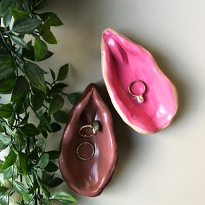 Könnte beinhalten: Zwei Keramik-Ringteller in Form einer Muschel. Eine Schale ist rosa und die andere braun. Beide Schalen enthalten goldene Ringe.