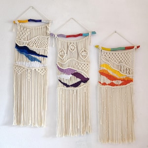 Può includere: Tre arazzi da parete in macramè con cotone color crema e accenti colorati. Ogni pezzo presenta un design unico con tonalità blu, viola e arancioni e una frangia nella parte inferiore. Gli arazzi sono sospesi a tasselli di legno.