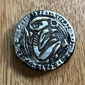 Könnte beinhalten: Ein runder, silberfarbener Anhänger mit schwarzem Rand. Der Anhänger zeigt eine stilisierte Skelettfigur in der Mitte, umgeben von einem verzierten, gravierten Design. Der Hintergrund ist eine Holzoberfläche.