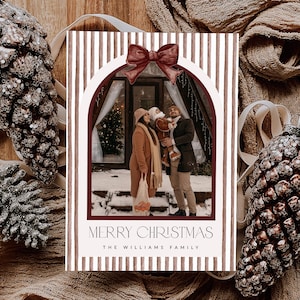 May include: A rectangular Christmas card featuring a vertical brown and white striped border with a dark red bow at the top. The center displays a photograph of a family in a snowy setting. The text reads MERRY CHRISTMAS and THE WILLIAMS FAMILY.