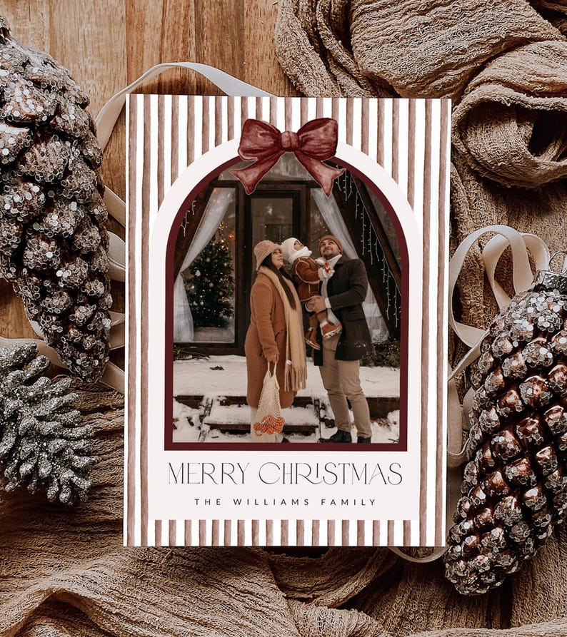 May include: A rectangular Christmas card featuring a vertical brown and white striped border with a dark red bow at the top. The center displays a photograph of a family in a snowy setting. The text reads MERRY CHRISTMAS and THE WILLIAMS FAMILY.