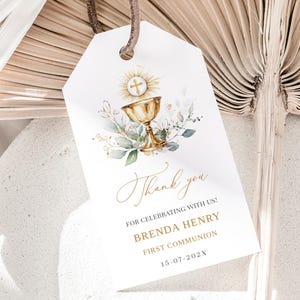 Puede incluir: Etiqueta blanca con una ilustración de cáliz dorado, cruz y detalles florales. La etiqueta dice "Thank you" en cursiva, con detalles de Primera Comunión. Una cuerda marrón está unida en la parte superior.