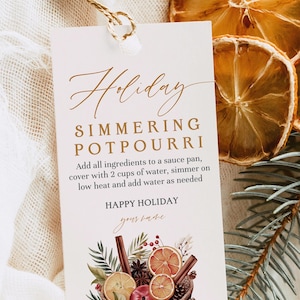 May include: A rectangular white paper tag with a gold string, featuring the text Holiday SIMMERING POTPOURRI, instructions to add ingredients to a sauce pan with 2 cups of water, and a graphic of dried orange slices, cinnamon sticks, and pine.