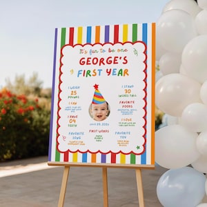 May include: A rectangular milestone birthday sign with a colorful vertical striped border. The text reads It's fun to be one, George's First Year, with sections for weight, height, teeth, favorite foods, songs, and toys, plus a photo of a baby.