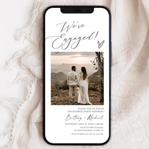 Könnte beinhalten: Ein Smartphone zeigt eine Verlobungsankündigung. Der Text "We're Engaged!" steht in eleganter Schrift über einem Foto eines Paares. Darunter eine Einladung mit Namen, Datum, Uhrzeit und Ort.