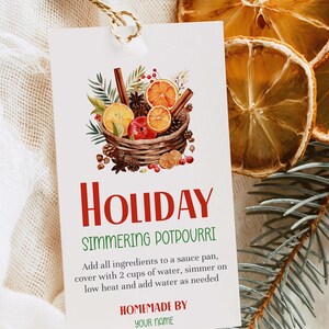 Könnte beinhalten: Ein rechteckiges weißes Etikett mit einer Schnurschlaufe, das eine Aquarellillustration eines geflochtenen Korbs mit Orangenscheiben, Zimtstangen, Tannenzapfen und Beeren zeigt. Der Text lautet Holiday Simmering Potpourri sowie Anweisungen und Homemade By Your Name.