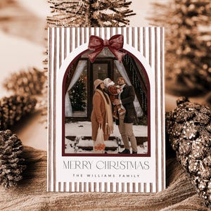 May include: A rectangular Christmas card featuring a vertical brown and white striped pattern with a dark red bow at the top of an arched photo frame. The photo shows a family of three standing in front of a cabin. Text reads MERRY CHRISTMAS THE WILLIAMS FAMILY.