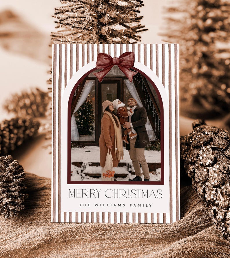 May include: A rectangular Christmas card featuring a vertical brown and white striped pattern with a dark red bow at the top of an arched photo frame. The photo shows a family of three standing in front of a cabin. Text reads MERRY CHRISTMAS THE WILLIAMS FAMILY.