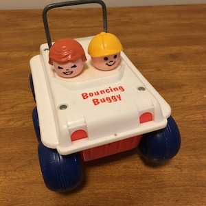 Puede incluir: Un coche de juguete vintage blanco y rojo con las palabras "Bouncing Buggy" impresas en la parte delantera. El juguete tiene dos figuras sonrientes y ruedas azules. Un asa gris se arquea sobre la parte superior del coche.