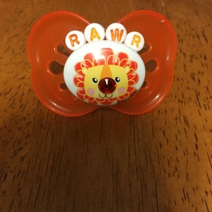May include: An orange baby pacifier with a lion design. The pacifier has the word "RAWR" in white letters above the lion's face. The lion has a red gem nose and a yellow and orange mane.