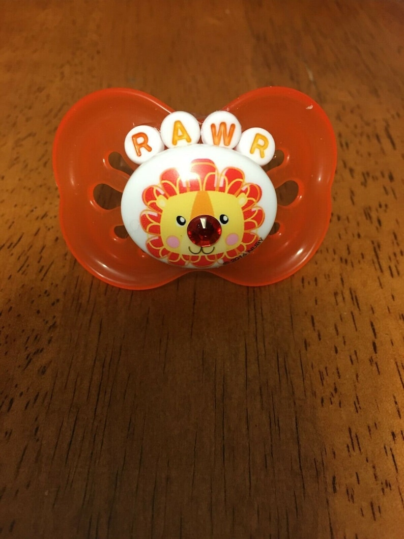 May include: An orange baby pacifier with a lion design. The pacifier has the word "RAWR" in white letters above the lion's face. The lion has a red gem nose and a yellow and orange mane.