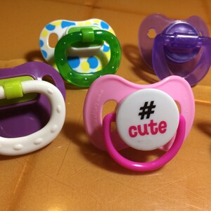 May include: A collection of colorful baby pacifiers in various shades of purple, pink, green, and white. One pacifier has the word "cute" printed on it. The pacifiers are made of plastic and have a round shape.