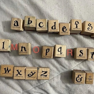 May include: A collection of wooden alphabet stamps featuring lowercase letters from a to z, an ampersand, and a heart symbol. Each square block displays a black serif typeface character on its top surface, arranged in three rows on a fabric surface.