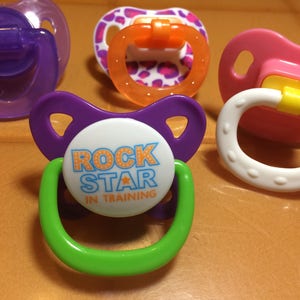 May include: A collection of colorful baby pacifiers. One pacifier has the text "ROCK STAR IN TRAINING" on the front. The pacifiers are in shades of purple, pink, orange, green, and white.