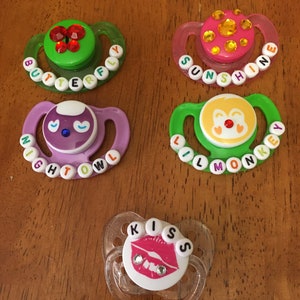 May include: Five baby pacifiers with decorative designs. The pacifiers have various colors, including green, pink, and purple. Each pacifier has a different design, such as a butterfly, the word "Sunshine", a night owl, a monkey, and the word "Kiss".