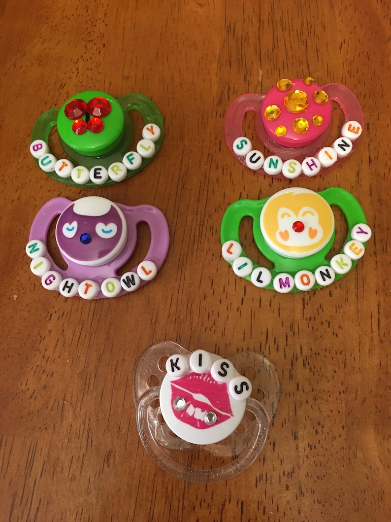 May include: Five baby pacifiers with decorative designs. The pacifiers have various colors, including green, pink, and purple. Each pacifier has a different design, such as a butterfly, the word "Sunshine", a night owl, a monkey, and the word "Kiss".