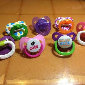 May include: A collection of colorful baby pacifiers. The pacifiers feature various designs, including polka dots, solid colors, and text. The colors include pink, purple, green, and orange. Some pacifiers have the text "# cute" and "ROCK STAR IN TRAINING".