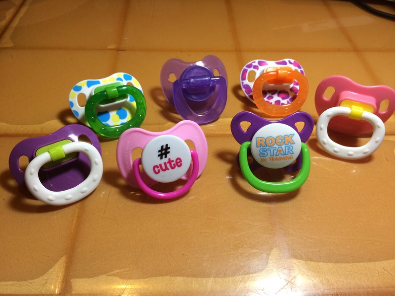 May include: A collection of colorful baby pacifiers. The pacifiers feature various designs, including polka dots, solid colors, and text. The colors include pink, purple, green, and orange. Some pacifiers have the text "# cute" and "ROCK STAR IN TRAINING".