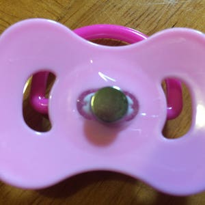May include: A pink baby pacifier with a round nipple and a shield-shaped mouth guard. The pacifier has a loop handle in a matching pink color. The nipple is a metallic color.