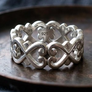 Puede incluir: Un anillo de plata de ley con un diseño calado con símbolos de corazón e infinito. El anillo está marcado con "925" y "09". El anillo se muestra sobre una superficie oscura y texturizada. El texto "Abdul Jewelrs" es visible.