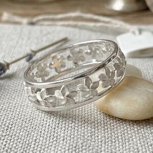 Può includere: Un anello d'argento con un motivo floreale. L'anello presenta una fascia di fiori ritagliati. È esposto su una superficie grigio chiaro con un rametto di lavanda e una piccola pietra. Il testo "Abdul Jewelrs" si trova nell'angolo in basso a destra.