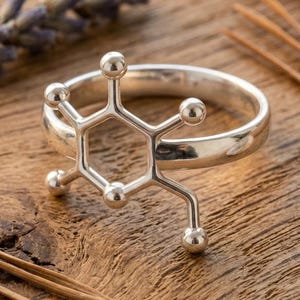 May include: A silver ring with a molecular structure design. The ring features a hexagonal shape with spherical accents. The ring is displayed on a wooden surface with dried lavender and other natural elements. The brand name "Abdul Jewelrs" is visible.
