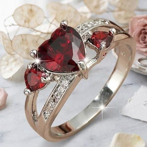 May include: A silver-toned ring with a large heart-shaped red gemstone and two smaller red gemstones. The ring is accented with small, clear gemstones. The ring is on a marble surface with flowers and the text "Beloved" and "Abdul Jewerls".