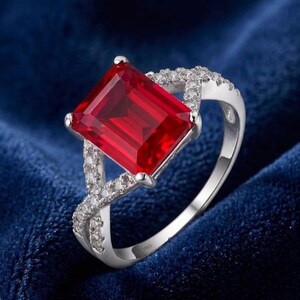 May include: A silver ring featuring a large, rectangular, vibrant red gemstone. The ring's band has a crisscross design with small, clear accent stones. The ring is set against a dark blue background.