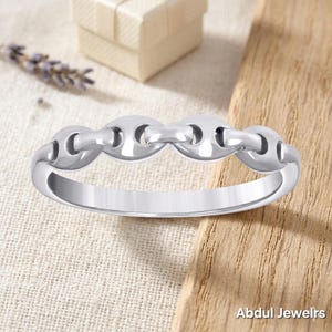 May include: A silver-colored ring with a chain link design. The ring is displayed on a wooden surface with a small gift box and lavender sprig in the background. The ring is a piece of jewelry.