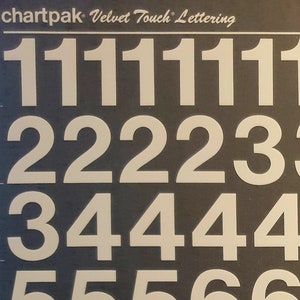 Peut inclure: Une feuille de lettres et de symboles autocollants blancs sur fond noir. Les lettres sont en police Helvetica et comprennent les chiffres de 0 à 9, des signes de ponctuation et des symboles monétaires. La feuille est étiquetée "chartpak Velvet Touch Lettering" et "helvetica medium 96PT./M 10096N".