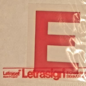 Op de afbeelding: Rode plastic letter E in Helvetica Medium lettertype, 50 mm groot. De letter is verpakt in een doorzichtige plastic zak met de merknaam "Letraset Letrasign" en de tekst "6 Tekens Stick/Un..."