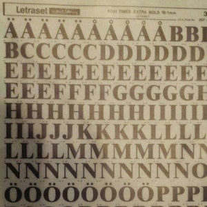 Puede incluir: Una hoja de letras de transferencia de fuente Letraset 6001 Times Extra Bold 16-1mm. La hoja incluye letras mayúsculas, números y símbolos. Las letras son negras y el fondo es crema.