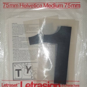 Op de afbeelding: Een close-up van een Letraset Letrasign-verpakking met vijf zwarte Helvetica Medium-lettertekens, elk 75 mm hoog. De verpakking bevat instructies voor het aanbrengen van de tekens.