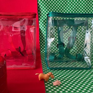 May include: Two clear plastic backpacks with red and teal trim. The backpacks have adjustable straps and are ideal for carrying small items.