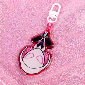 May include: A pink and white acrylic keychain featuring a cartoon character with a white mask and black eyes. The character is hanging upside down from a web.