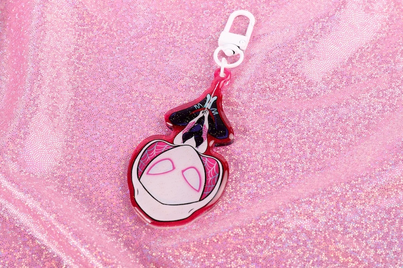 May include: A pink and white acrylic keychain featuring a cartoon character with a white mask and black eyes. The character is hanging upside down from a web.