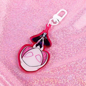May include: A pink and white acrylic keychain featuring a cartoon character with a white mask and black eyes. The character is hanging upside down from a web.