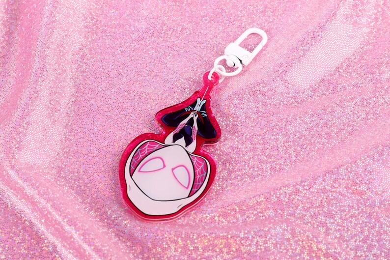 May include: A pink and white acrylic keychain featuring a cartoon character with a white mask and black eyes. The character is hanging upside down from a web.