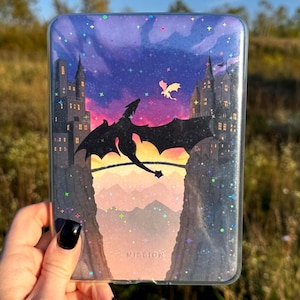 May include: A clear protective e-reader case featuring a printed graphic of a large black dragon flying between two stone castles at sunset. The sky is a gradient of purple and orange with holographic star accents and the word MISSION printed below.