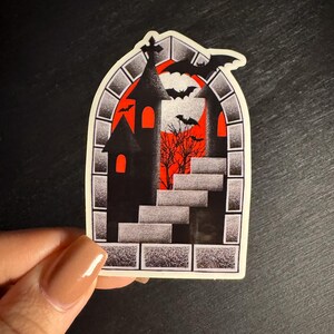 Puede incluir: Una pegatina de Halloween con una silueta de castillo negro, murci&eacute;lagos y una escalera sobre un fondo rojo y naranja. El dise&ntilde;o est&aacute; enmarcado por un borde de piedra. La pegatina est&aacute; sujeta por una mano.