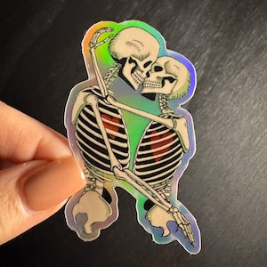 May include: Holographic sticker featuring two skeletons embracing and kissing. The skeletons are depicted in a detailed, anatomical style with visible rib cages and skulls. The sticker has a rainbow iridescent effect, creating a colorful sheen.