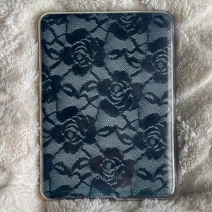May include: A rectangular, clear phone case featuring a black lace rose pattern. The case has a transparent border and is set against a fluffy, white background. The lace design is detailed and covers the entire back of the case.