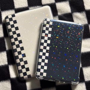 May include: Two rectangular protective cases for electronic devices. One case is white with a black and white checkered side panel and holographic star accents. The second case is black with a black and white checkered side panel and colorful holographic star patterns.