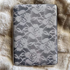 May include: A rectangular phone case with a white lace rose design on a grey backdrop. The case has rounded corners and a glossy finish. The word "SSION" is visible at the bottom of the case.