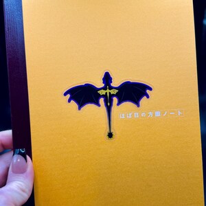 May include: A mustard-yellow notebook with a dark blue and gold dragon design. The dragon has outstretched wings and a long tail. Japanese text is printed on the upper right corner of the notebook.